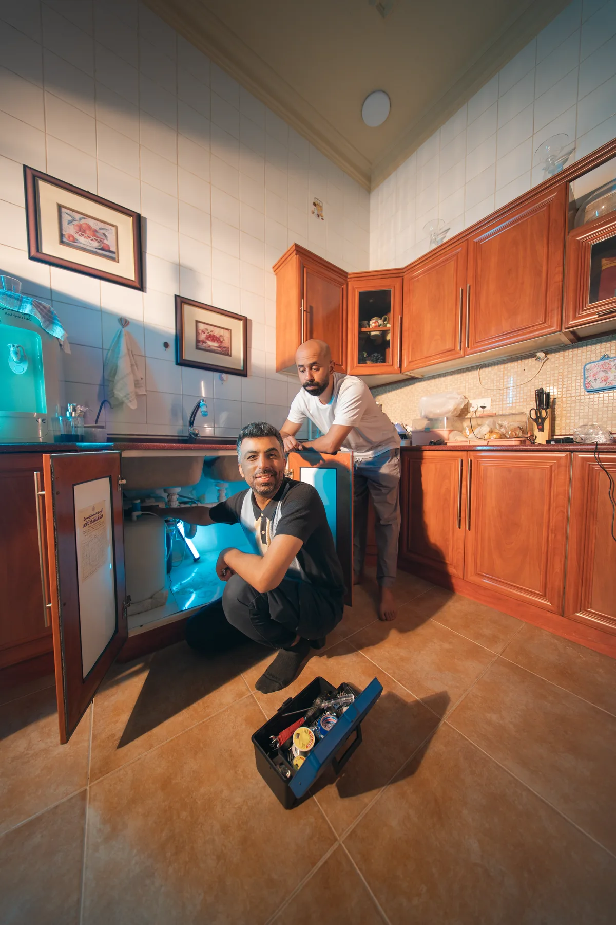 The plumber crouched by an open sink cabinet with Fahad leaning on the door behind him, FLOOSS campaign still